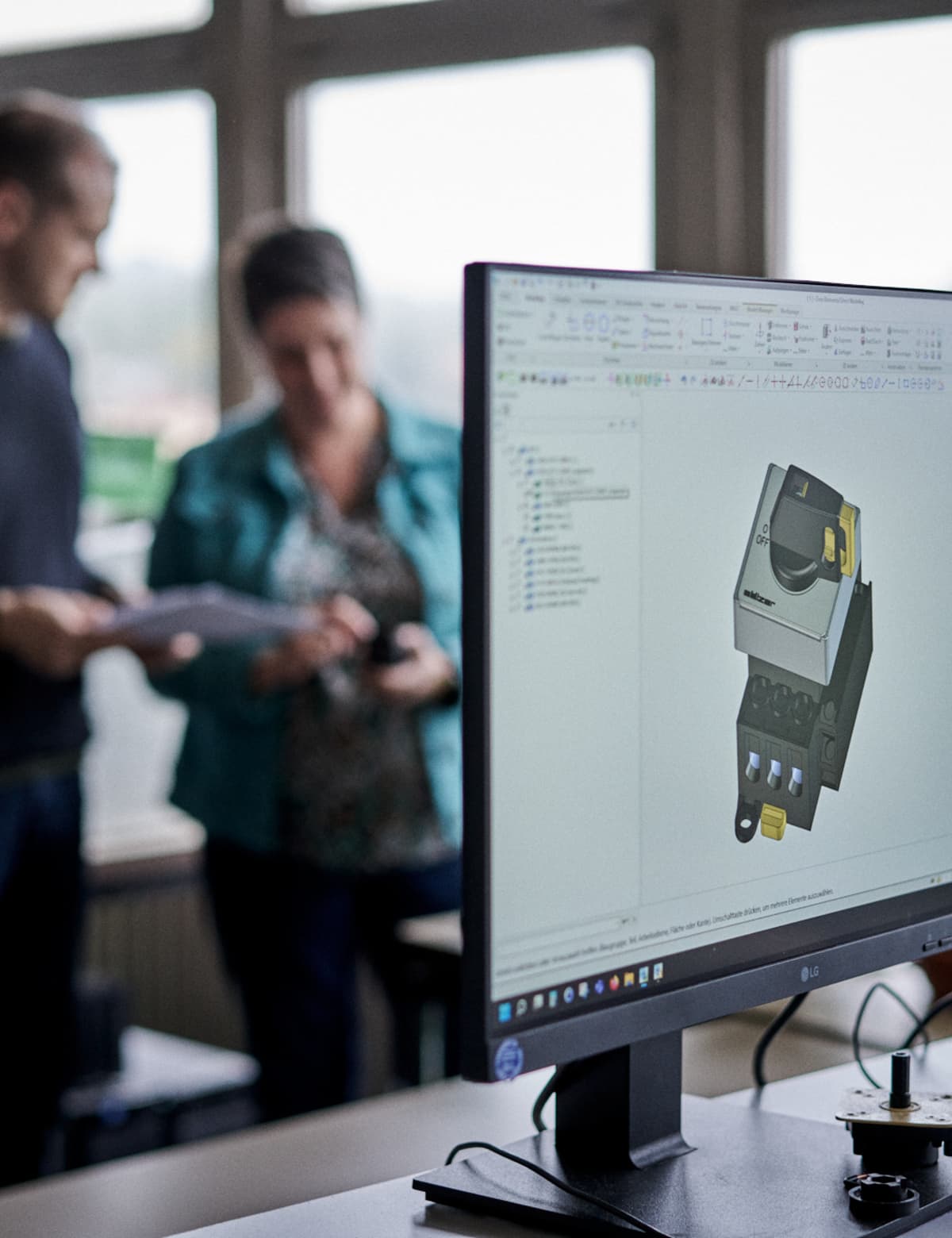 Zwei Personen, die einen Katalog durchblättern, währen im Vordergrund ein Monitor mit einem 3D-Modell eines Schalters zu sehen ist. 