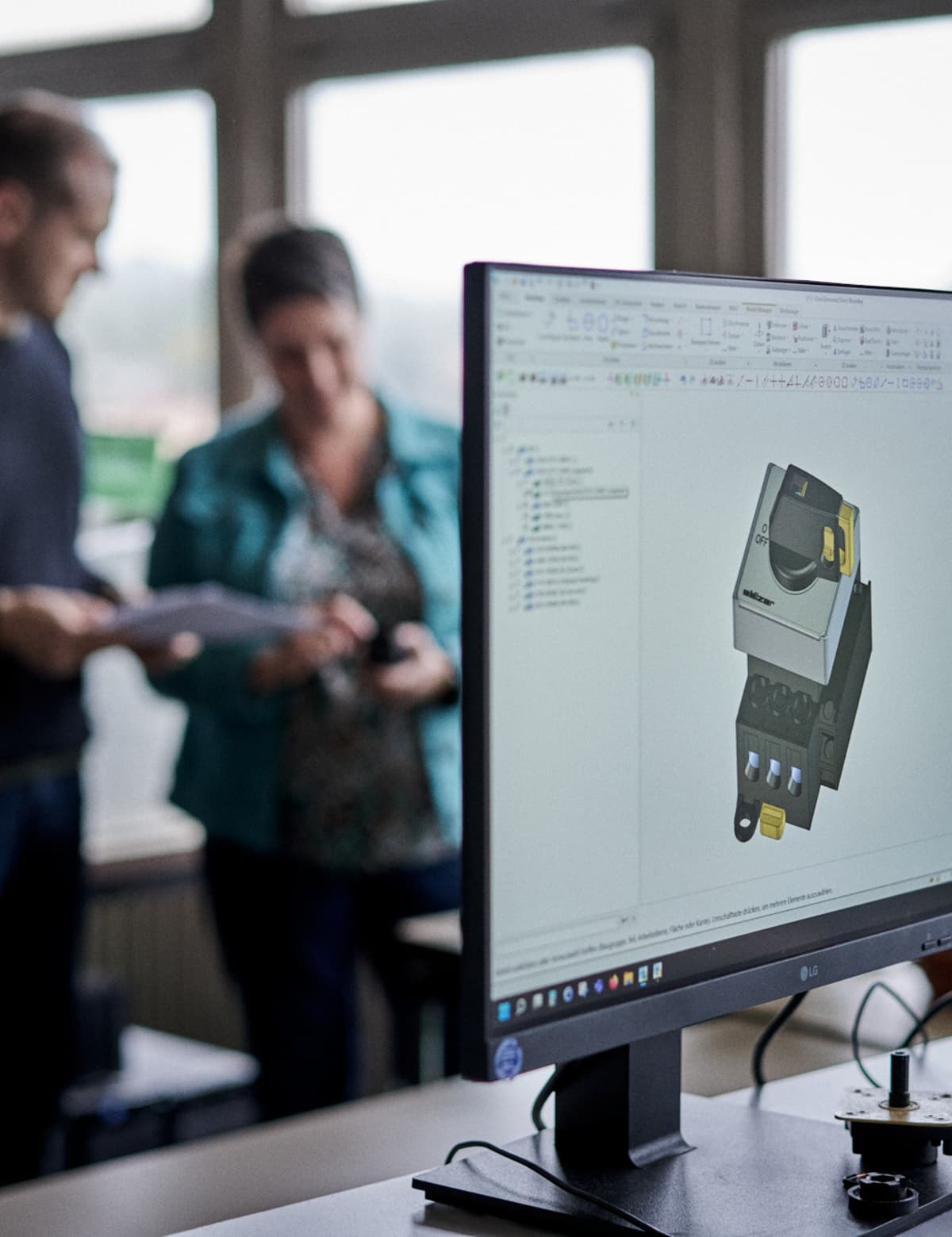 Zwei Personen, die einen Katalog durchblättern, währen im Vordergrund ein Monitor mit einem 3D-Modell eines Schalters zu sehen ist. 