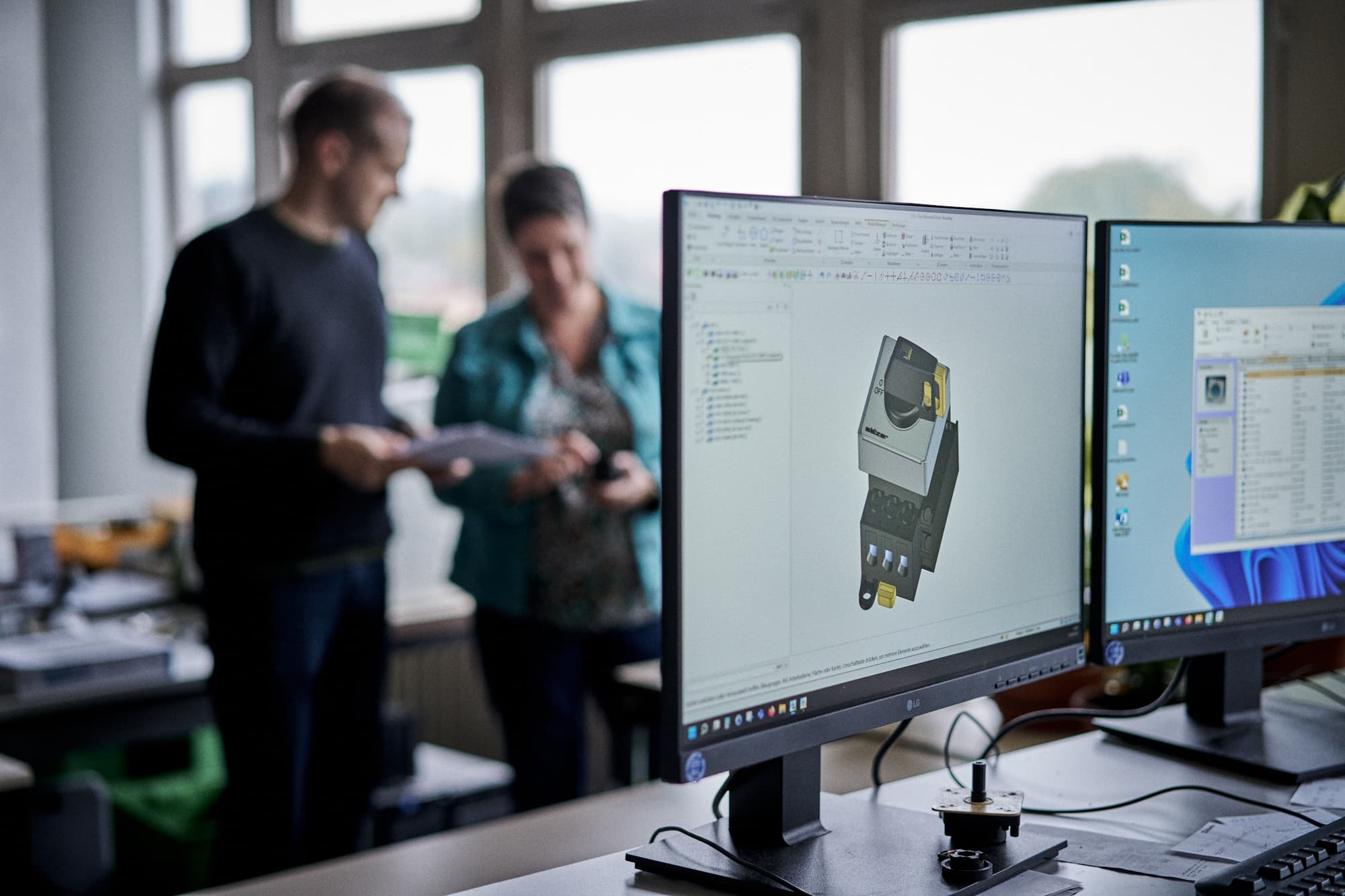 Zwei Personen, die einen Katalog durchblättern, währen im Vordergrund ein Monitor mit einem 3D-Modell eines Schalters zu sehen ist. 